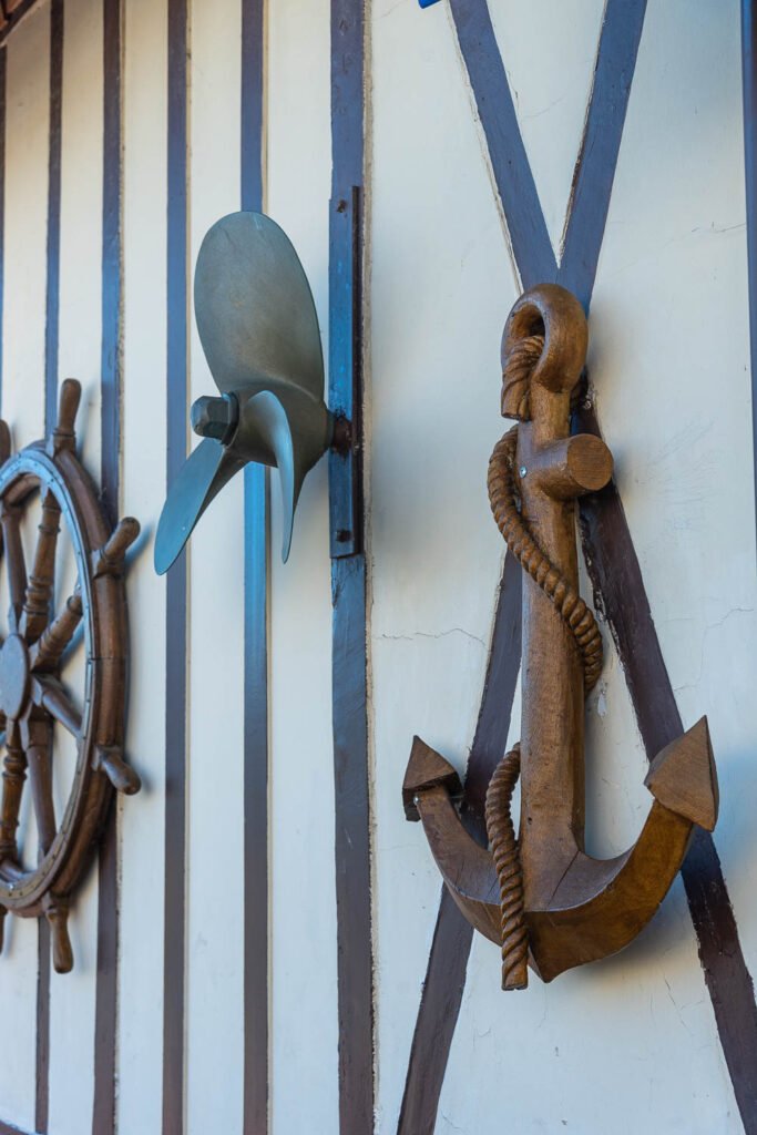 Nautical wall display with a large wooden ship wheel, an iron anchor bound by rope, and a metal propeller mounted on a striped wall.